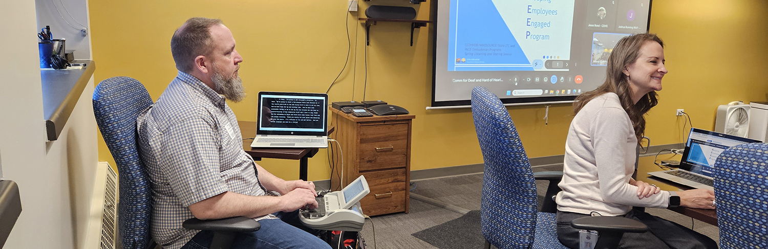 Male captioner sitting behind a presenter in a meeting room to provide captions via CART