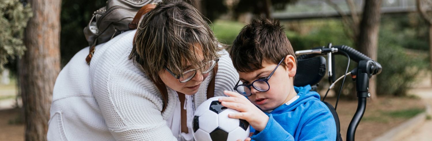 Adult woman  with short brown hair and glasses holding a soccer ball for a young boy with glasses in a wheelchair to touch