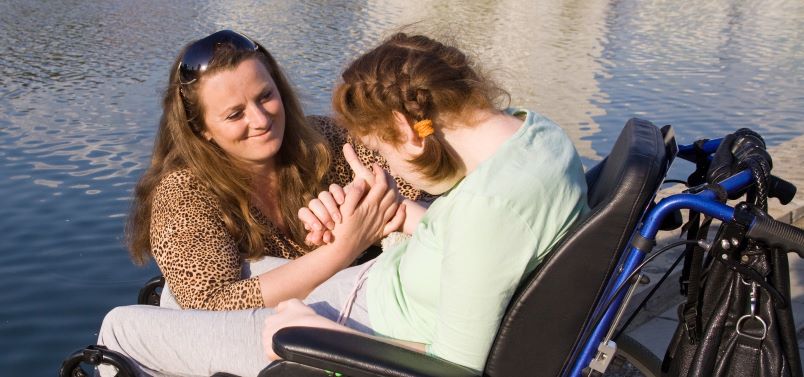 Smiling woman with long brown hair crouches next to a wheelchair and offers her hand to the young girl in the chair