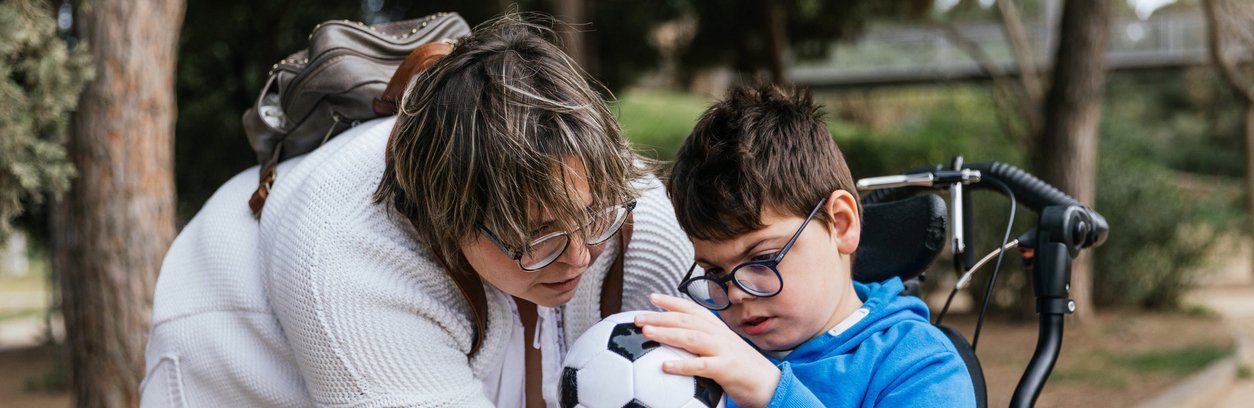 Adult woman with short brown hair and glasses holding a soccer ball for a young boy with glasses in a wheelchair to touch