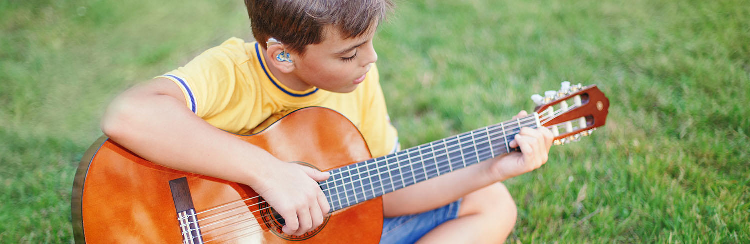 young boy with hearing aids plays guitar while sitting in the grass