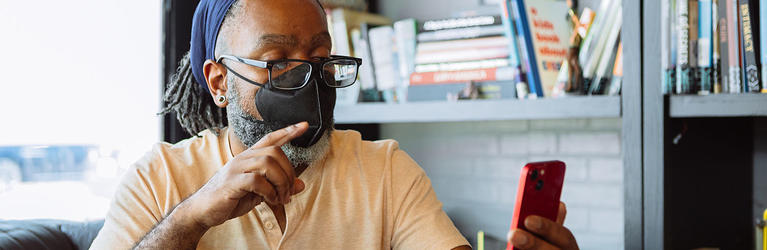 A Deaf Black man video chats via phone, signing with one hand while extending and tilting a smartphone back with the other hand. The man is in front of a bright window and has glasses, a KN-95 mask, and hair pulled back with a bandana.